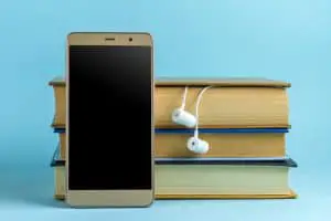 藍色背景上的耳機、手機和書籍。 有聲讀物的概念。 播放文學、教育書籍的音頻播放。
